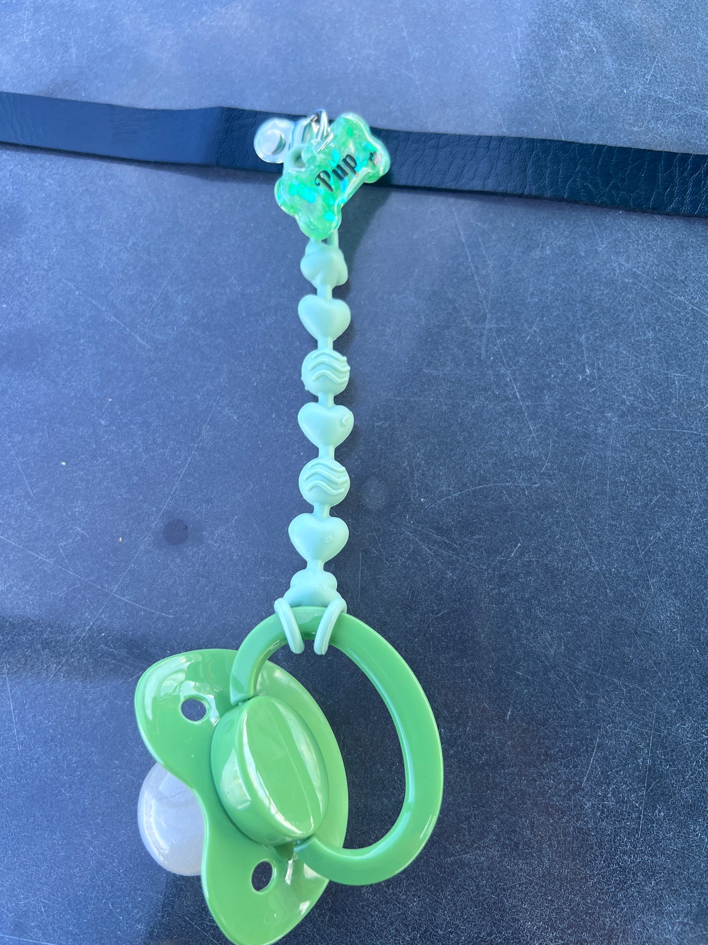 Adult pacifier choker featuring a comfortable choker with an attached adult pacifier, connected by a chewable silicone string. A customizable resin name tag is displayed at the center for personalization.