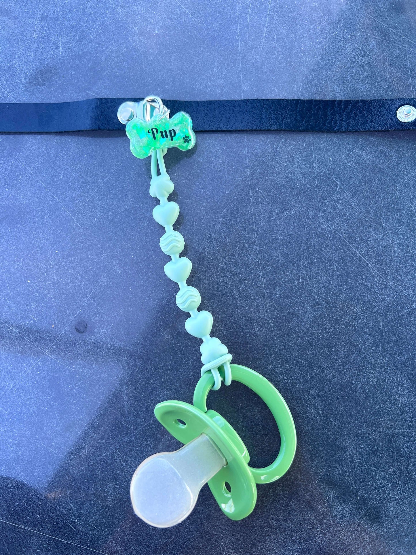 Adult pacifier choker featuring a comfortable choker with an attached adult pacifier, connected by a chewable silicone string. A customizable resin name tag is displayed at the center for personalization.