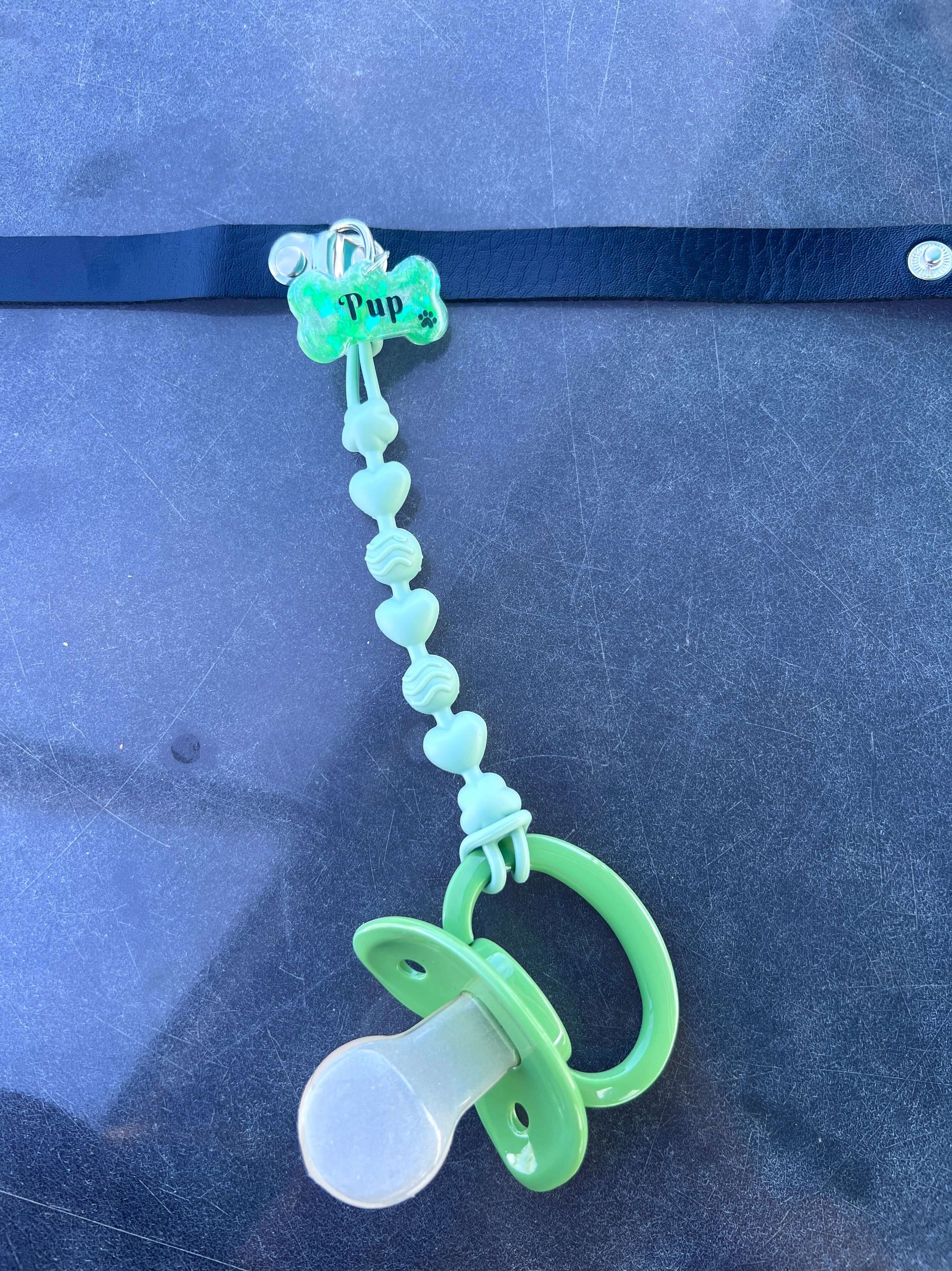 Adult pacifier choker featuring a comfortable choker with an attached adult pacifier, connected by a chewable silicone string. A customizable resin name tag is displayed at the center for personalization.