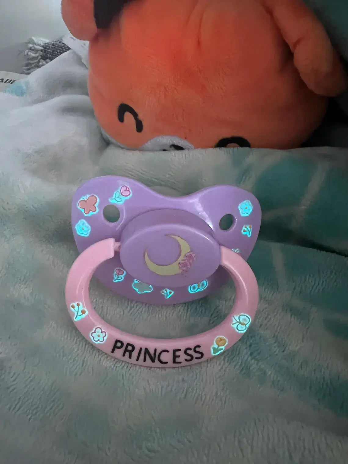 Adult deco pacifier featuring glow-in-the-dark flowers and butterflies on the shield, a moon centerpiece, and the word 'Princess' on the handle for a magical and elegant design.