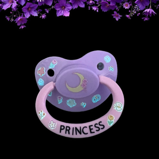 Adult deco pacifier featuring glow-in-the-dark flowers on the shield, a moon centerpiece, and the word 'Princess' on the handle for a magical and elegant design.