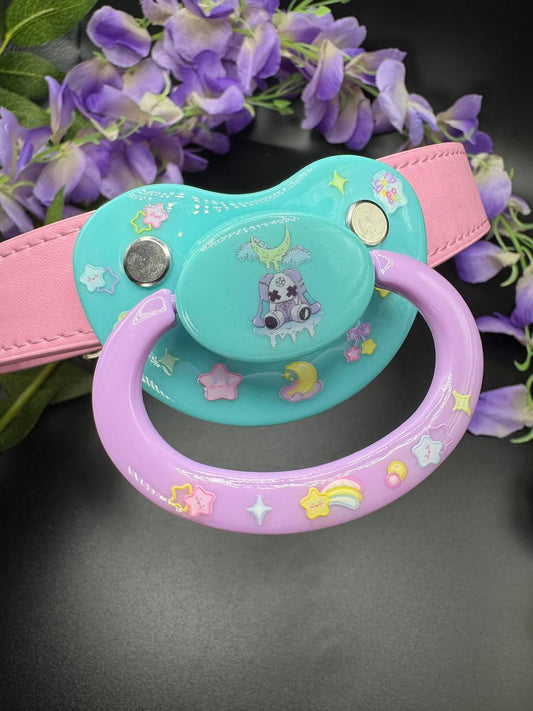 Aqua and lavender pacifier with pink gag straps. Showing a creepy kawaii bunny center with stars and moons that glow in the dark
