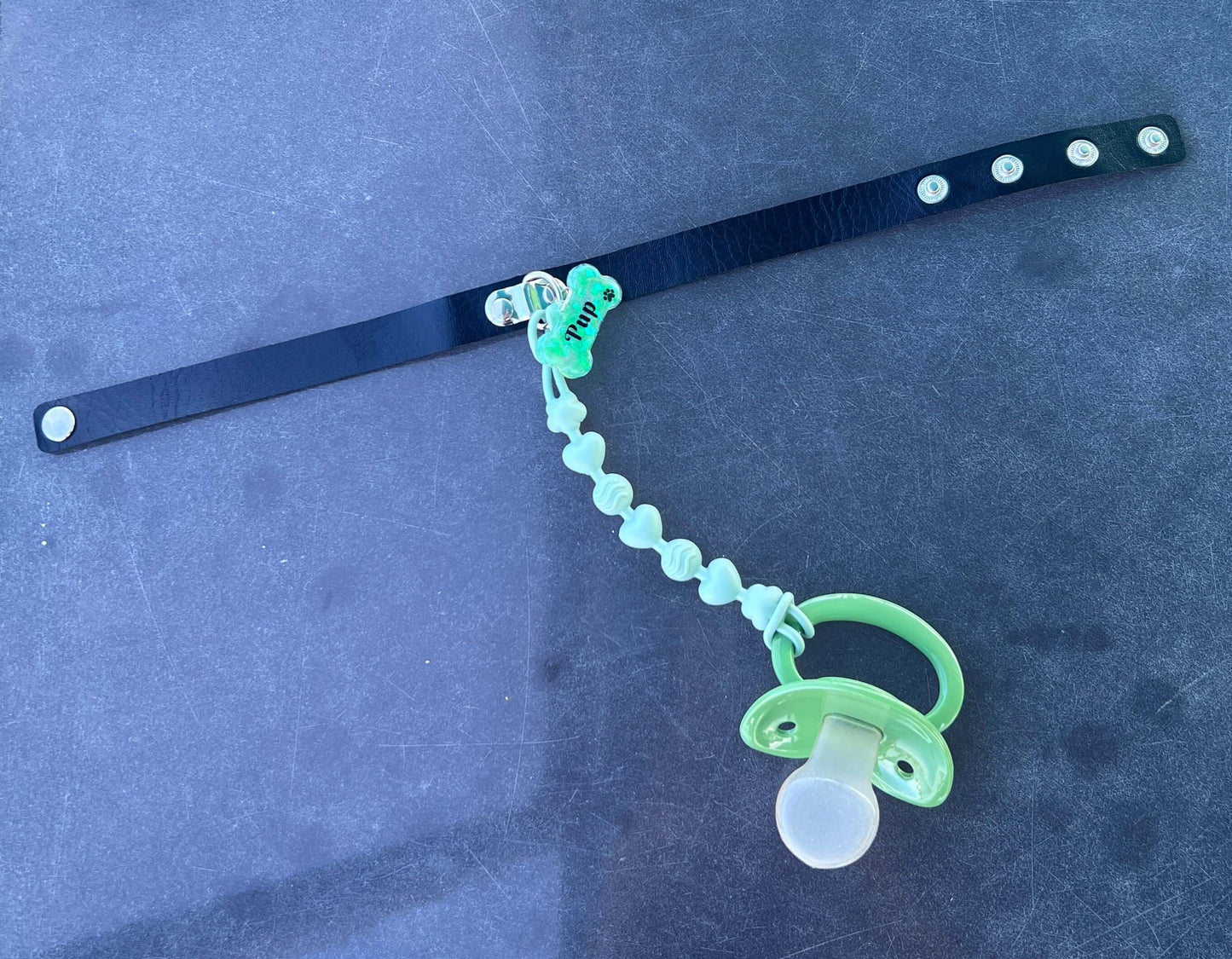 Adult pacifier choker featuring a comfortable choker with an attached adult pacifier, connected by a chewable silicone string. A customizable resin name tag is displayed at the center for personalization.