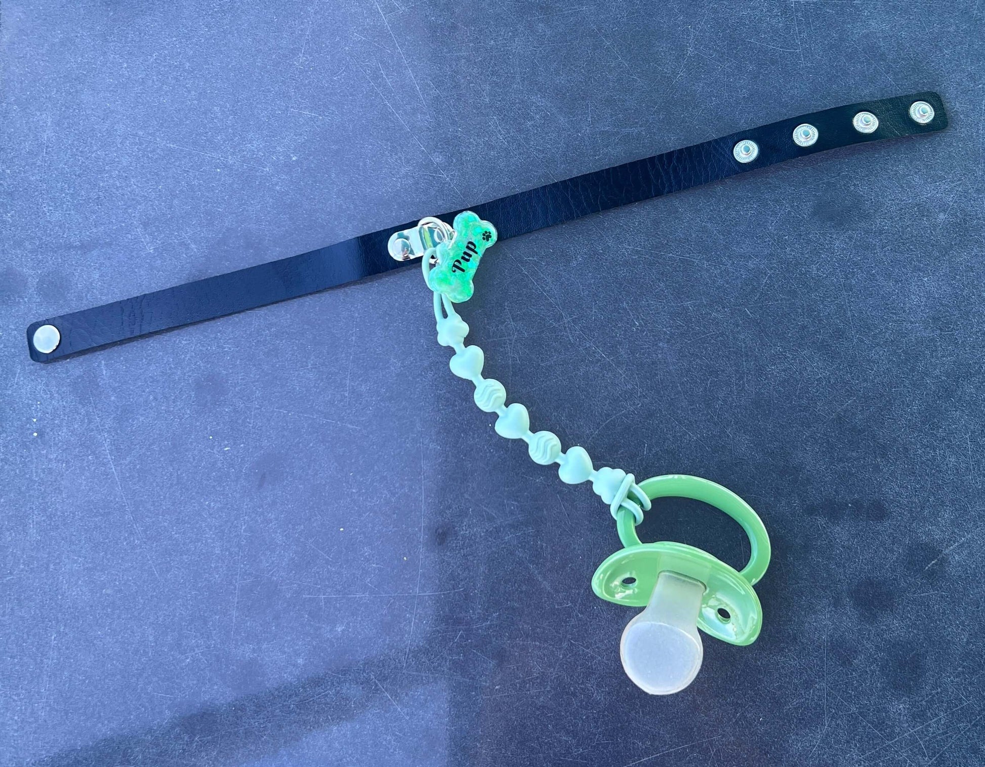 Adult pacifier choker featuring a comfortable choker with an attached adult pacifier, connected by a chewable silicone string. A customizable resin name tag is displayed at the center for personalization.