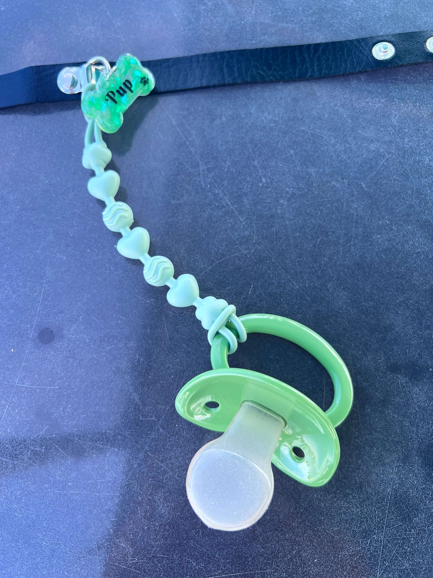 Adult pacifier choker featuring a comfortable choker with an attached adult pacifier, connected by a chewable silicone string. A customizable resin name tag is displayed at the center for personalization.