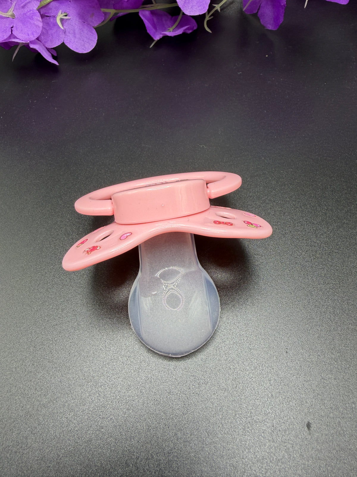 A pink adult pacifier with a translucent frosted nipple, resting on a dark gray surface with purple flowers in the background.