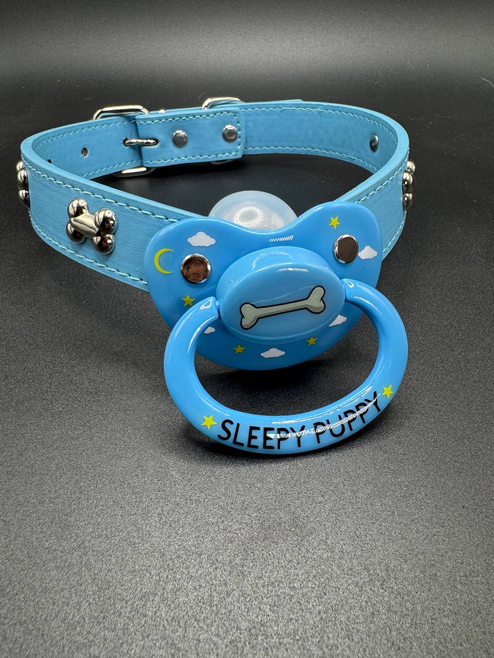 Blue dog collar straps with pacifier attachment that says Sleepy puppy with dog bone center and clouds and stars on shield on a gray background