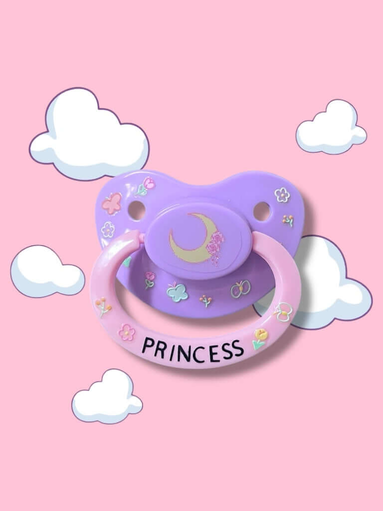 Adult deco pacifier featuring glow-in-the-dark flowers and butterflies on the shield, a moon centerpiece, and the word 'Princess' on the handle for a magical and elegant design.