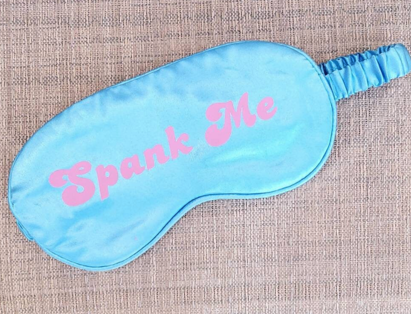 A blue silk adult blindfold with the text 'Spank Me' printed in pink on the front, featuring an elastic strap.