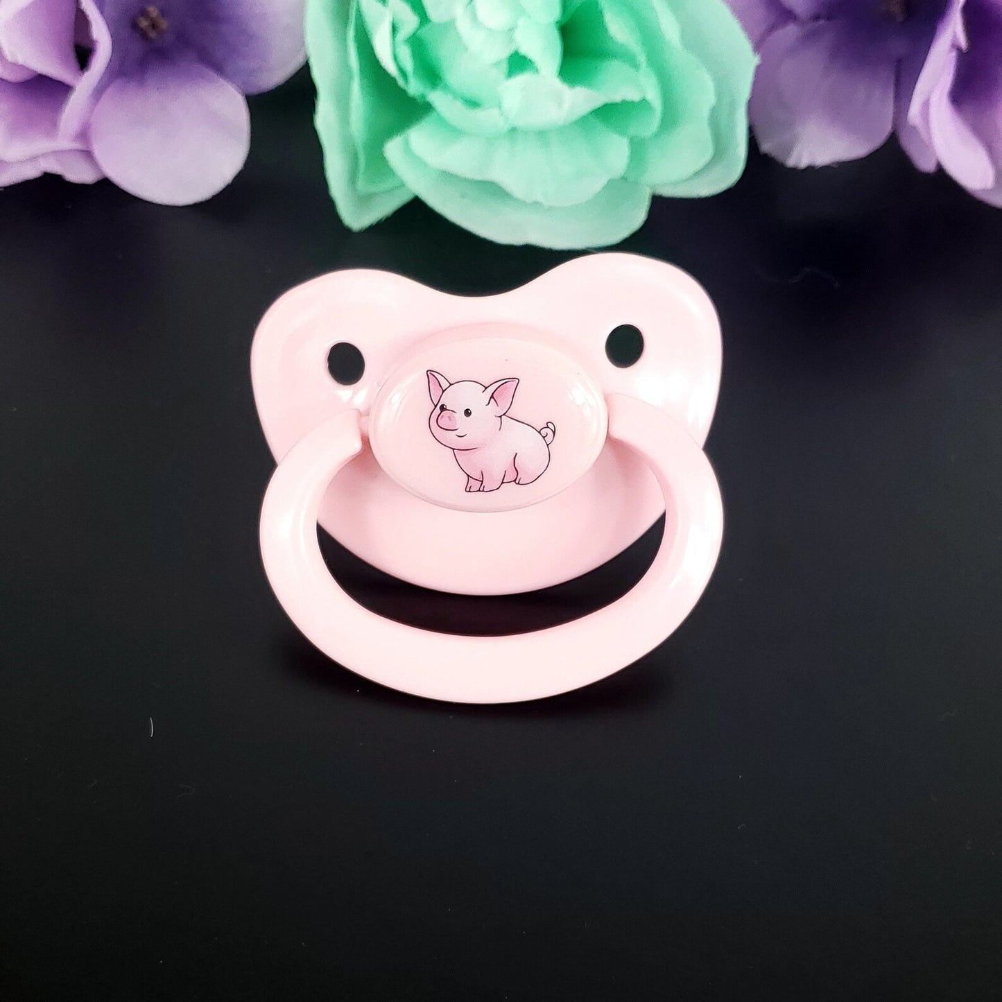 Pink adult pacifier with a pig illustration on a black background with flowers.