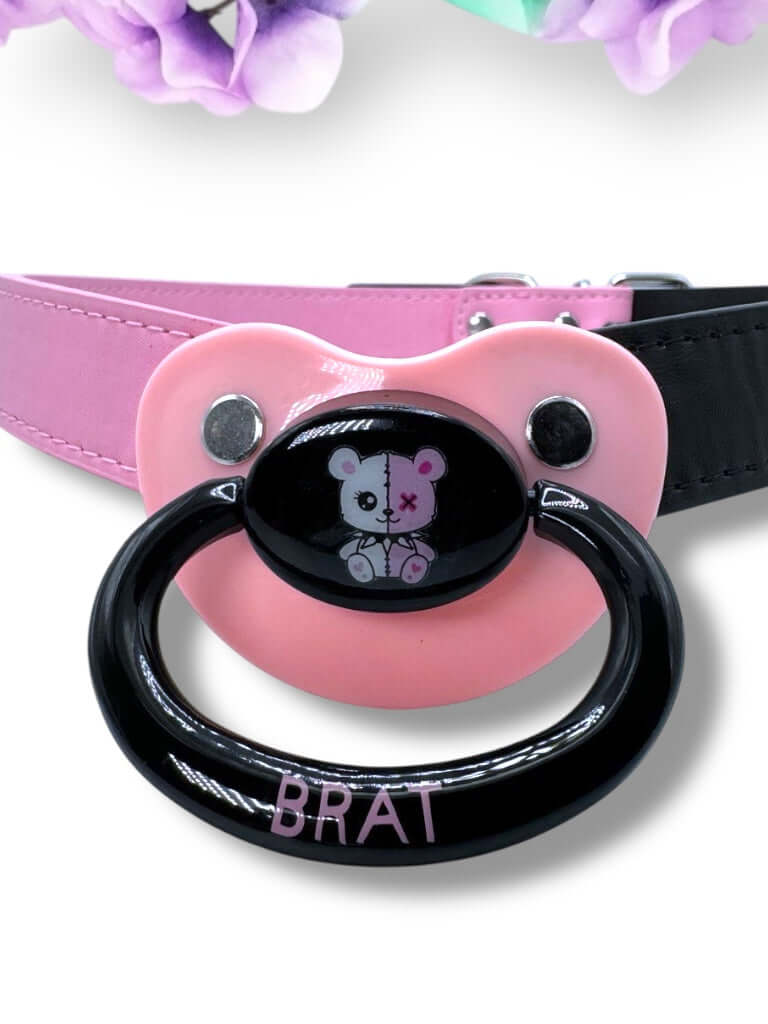 Adult pacifier gag featuring a black and pink pacifier with a punk bear design on the center button, the word 'Brat' in pink on the handle, and mismatched straps—one black and one pink—for a bold, edgy look.