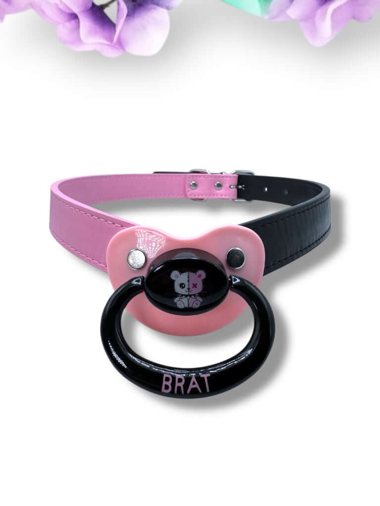 Adult pacifier gag featuring a black and pink pacifier with a punk bear design on the center button, the word 'Brat' in pink on the handle, and mismatched straps—one black and one pink—for a bold, edgy look.