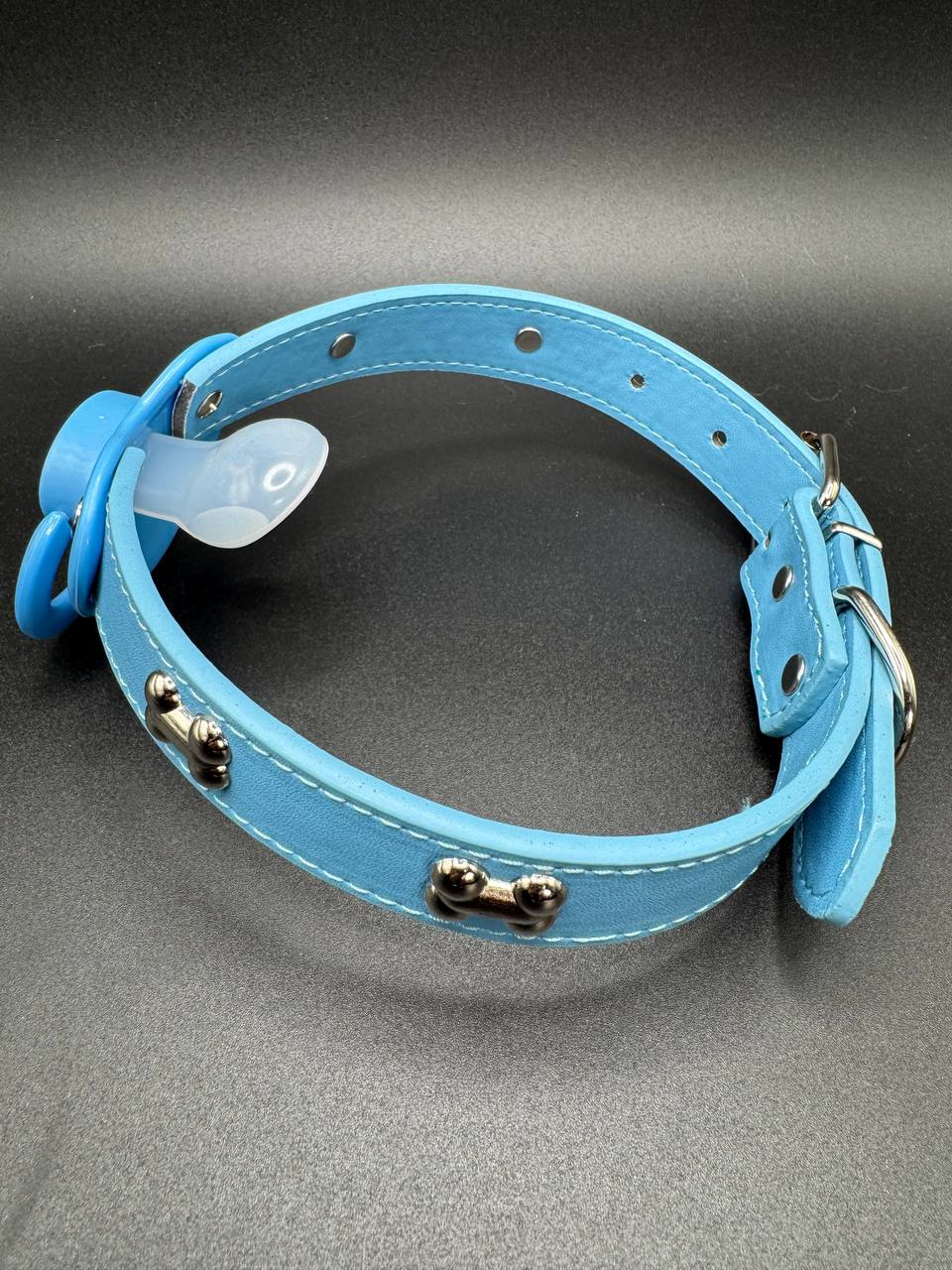 Blue leather pacifier gag straps with metal dog bone studs on a gray background