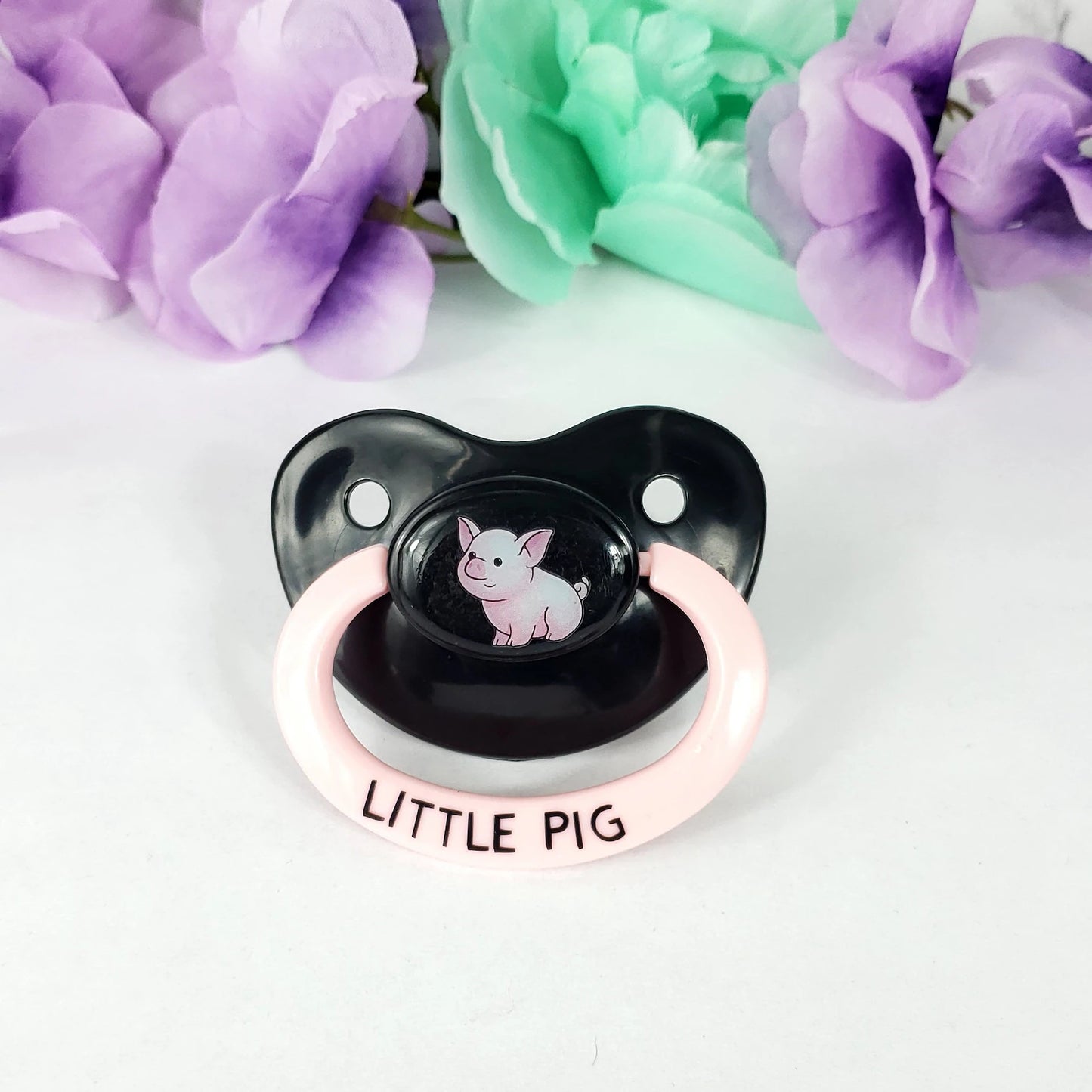 Black and pink pacifier with 'Little Pig' text and pig illustration, surrounded by flowers on a white background