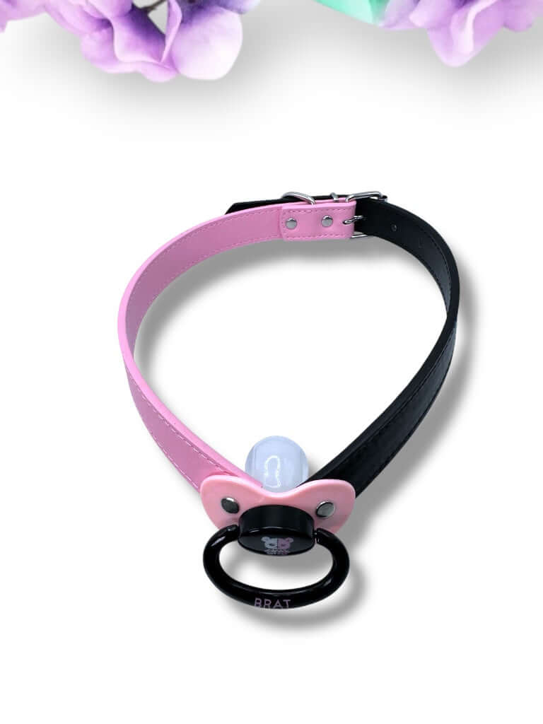 Adult pacifier gag featuring a black and pink pacifier with a punk bear design on the center button, the word 'Brat' in pink on the handle, and mismatched straps—one black and one pink—for a bold, edgy look.