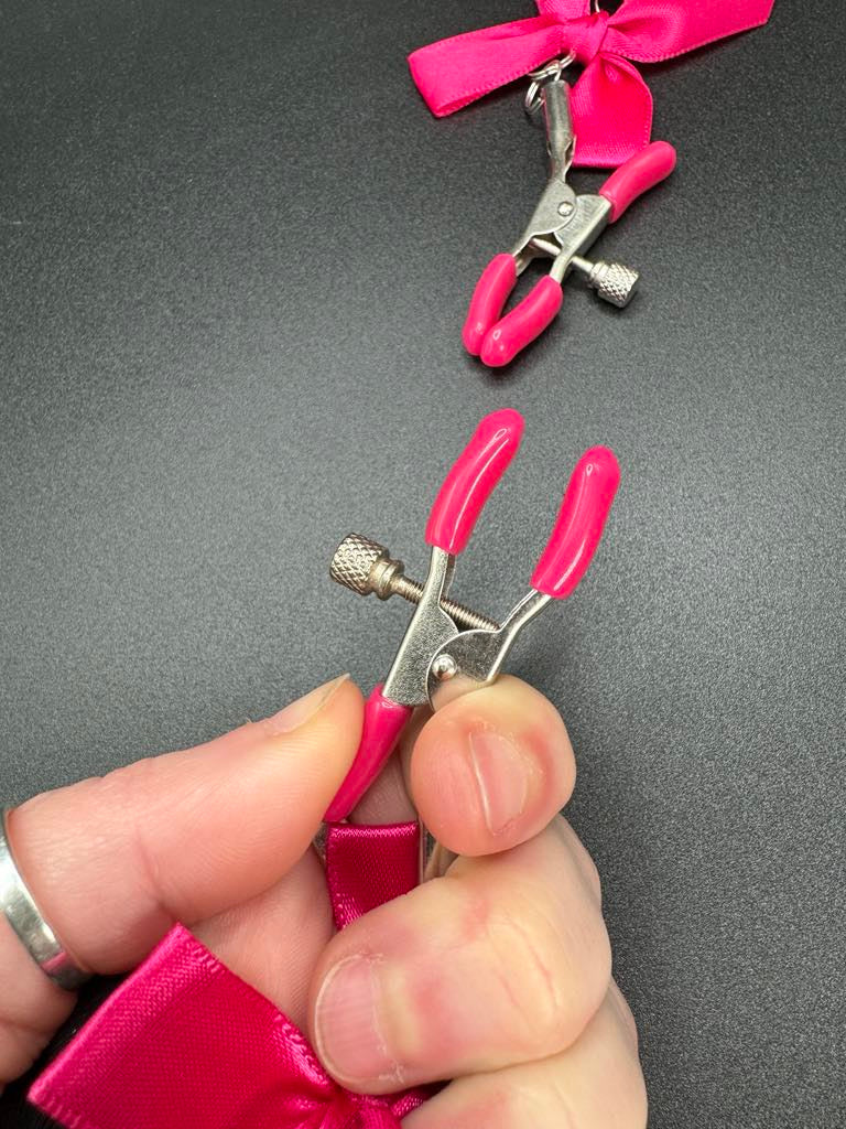 Stainless steel nipple clamps with attached bows and bells at the ends, featuring a silicone cover for added comfort and a stylish, playful touch.