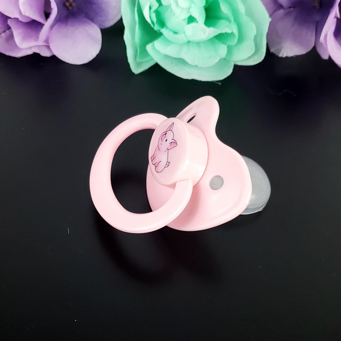 side view of Pink adult pacifier with a pig design on a black background with flowers