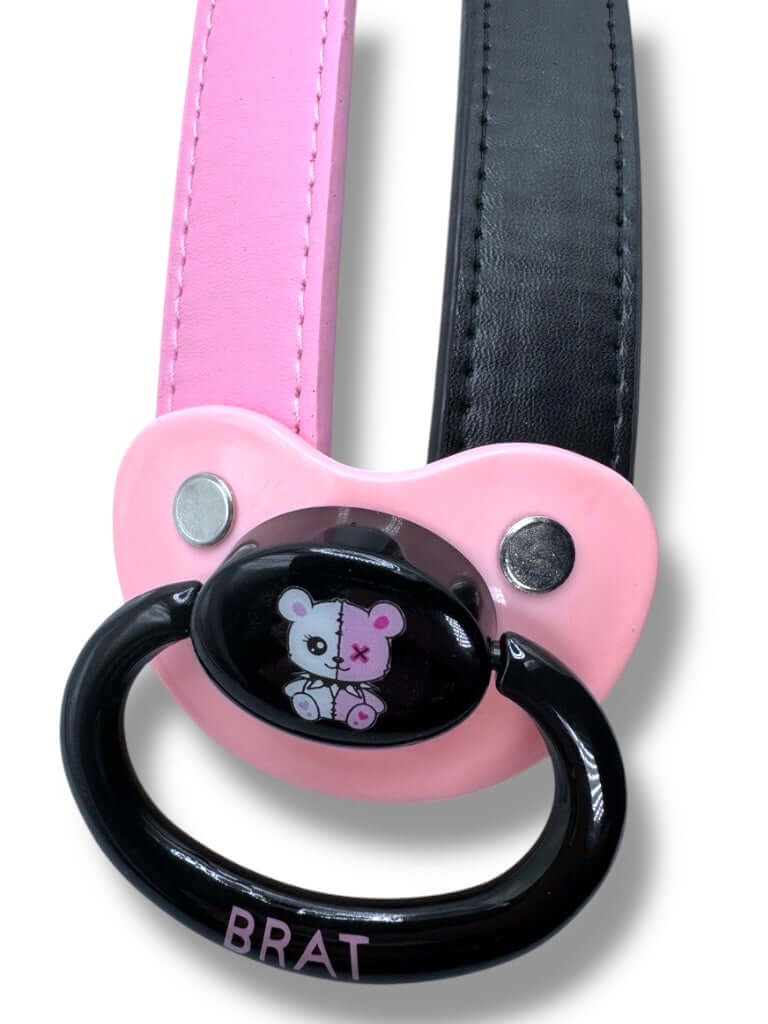 Adult pacifier gag featuring a black and pink pacifier with a punk bear design on the center button, the word 'Brat' in pink on the handle, and mismatched straps—one black and one pink—for a bold, edgy look.