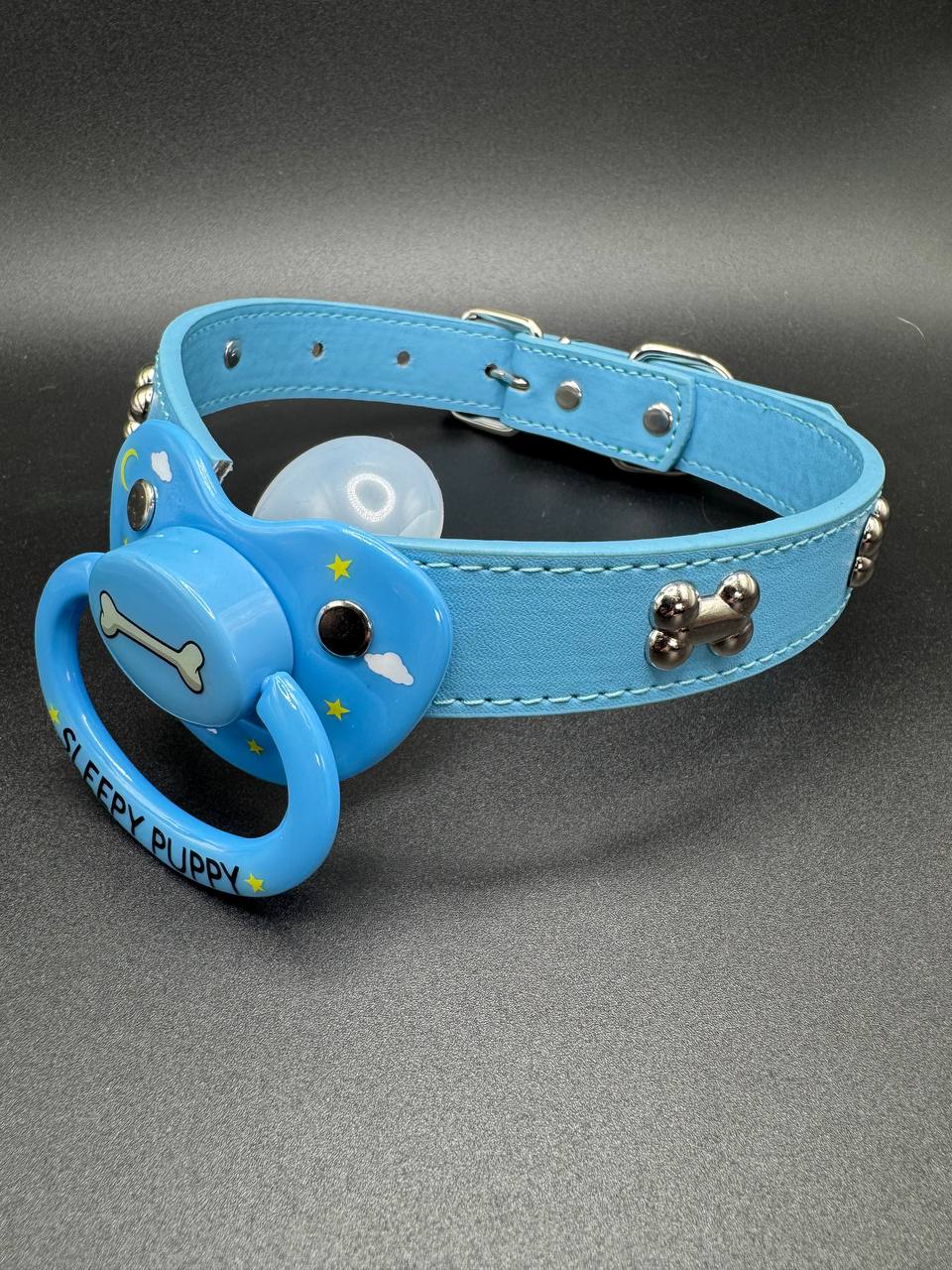 Blue dog bone collar with a pacifier attachment that is decorated with stars, clouds and a dog bone center with the text sleepy puppy on the handle on a gray background