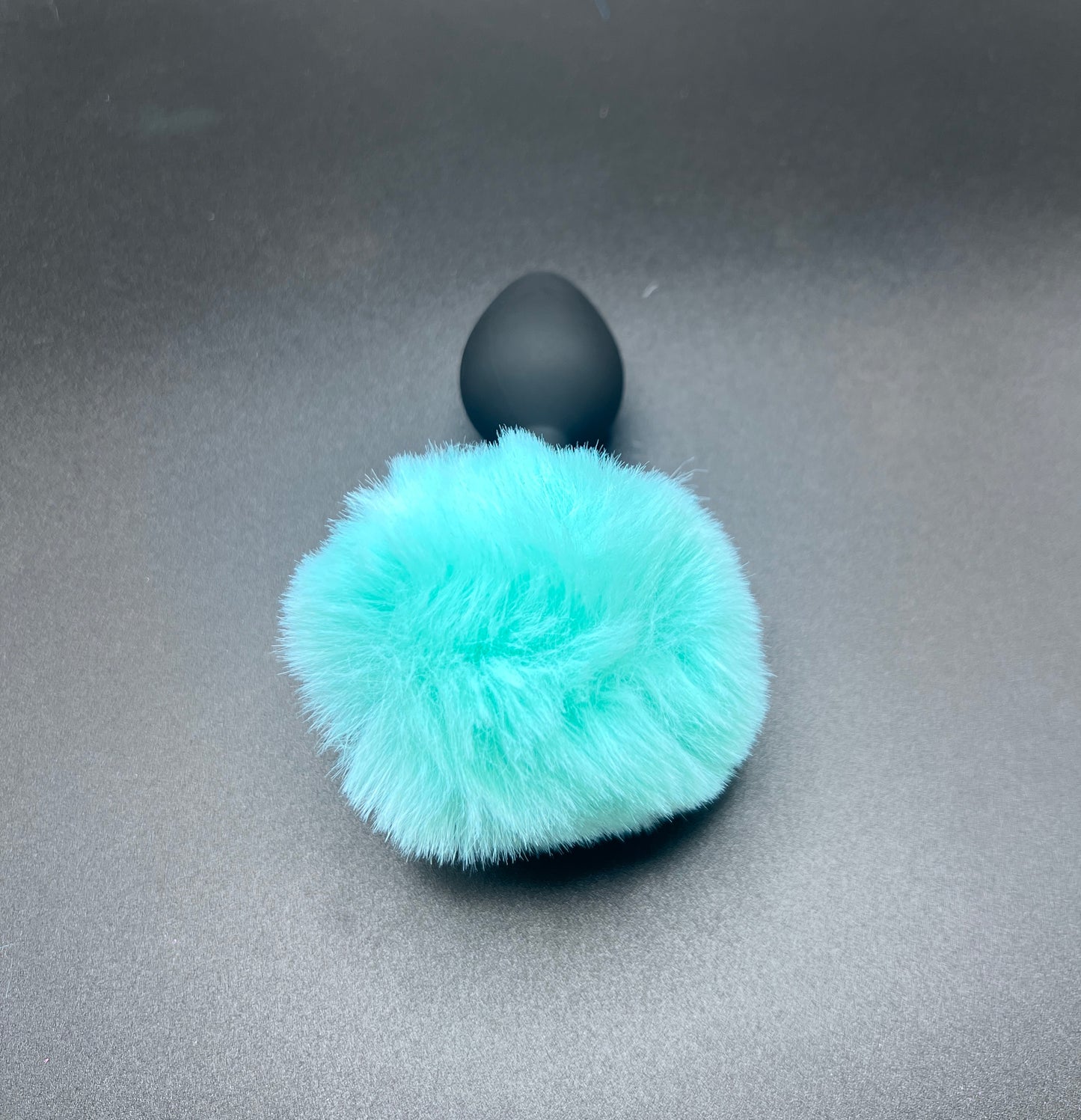 Black sex toy with a turquoise fur pom-pom bunny tail on a gray background