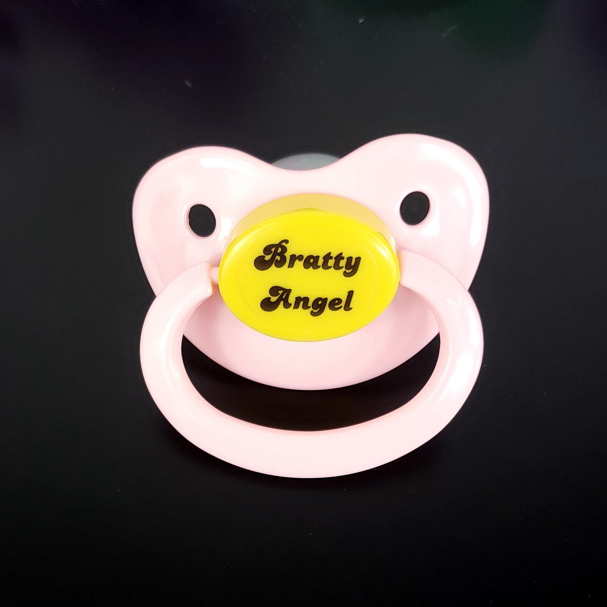 An adult pacifier with a soft pastel design featuring the phrase "Bratty Angel" in elegant text at the center, blending a playful and sweet aesthetic.