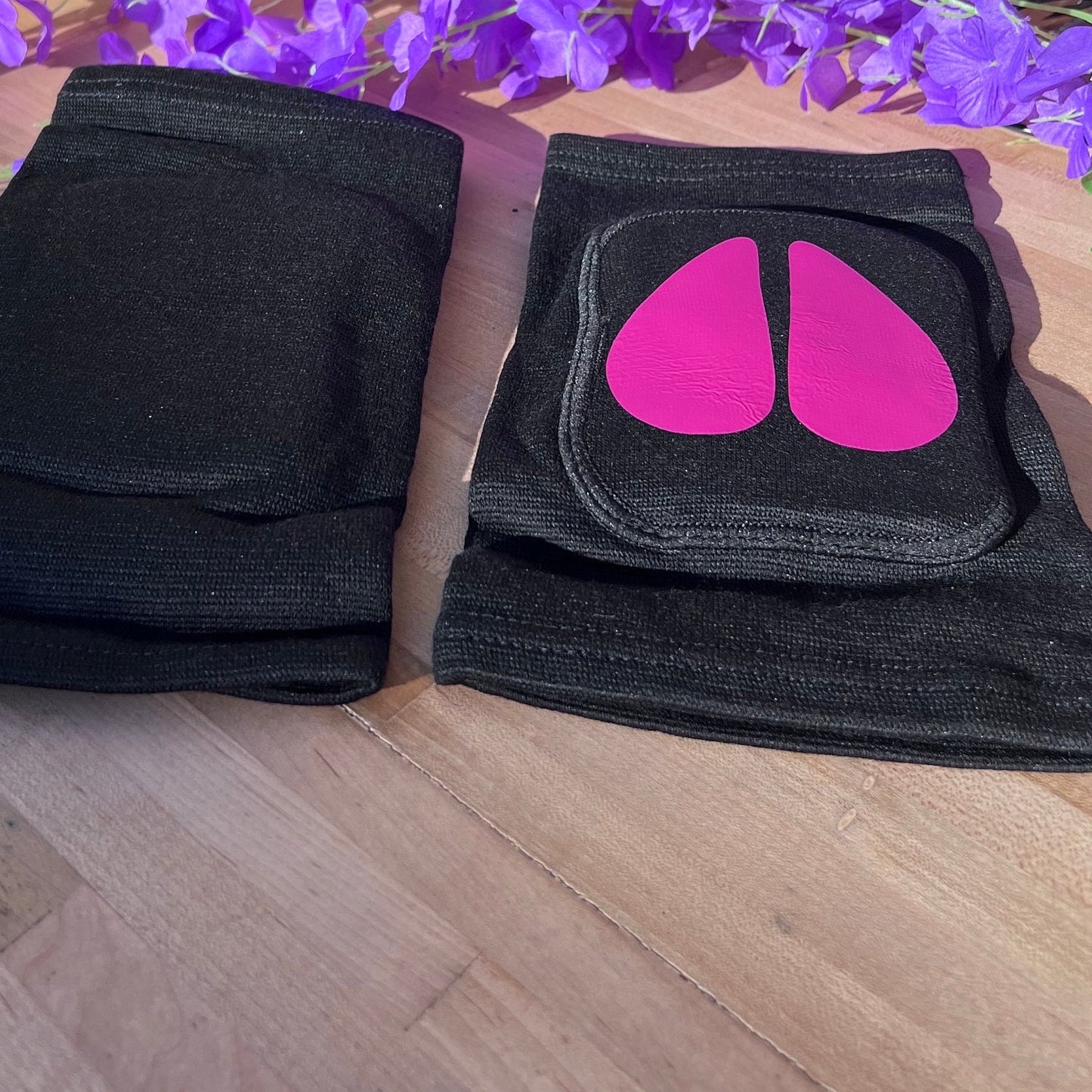 Another angle of the same black adult knee pads with bright pink cow hoof vinyl designs, one facing down, shown on a wooden surface with a bunch of purple artificial flowers arranged above them.
