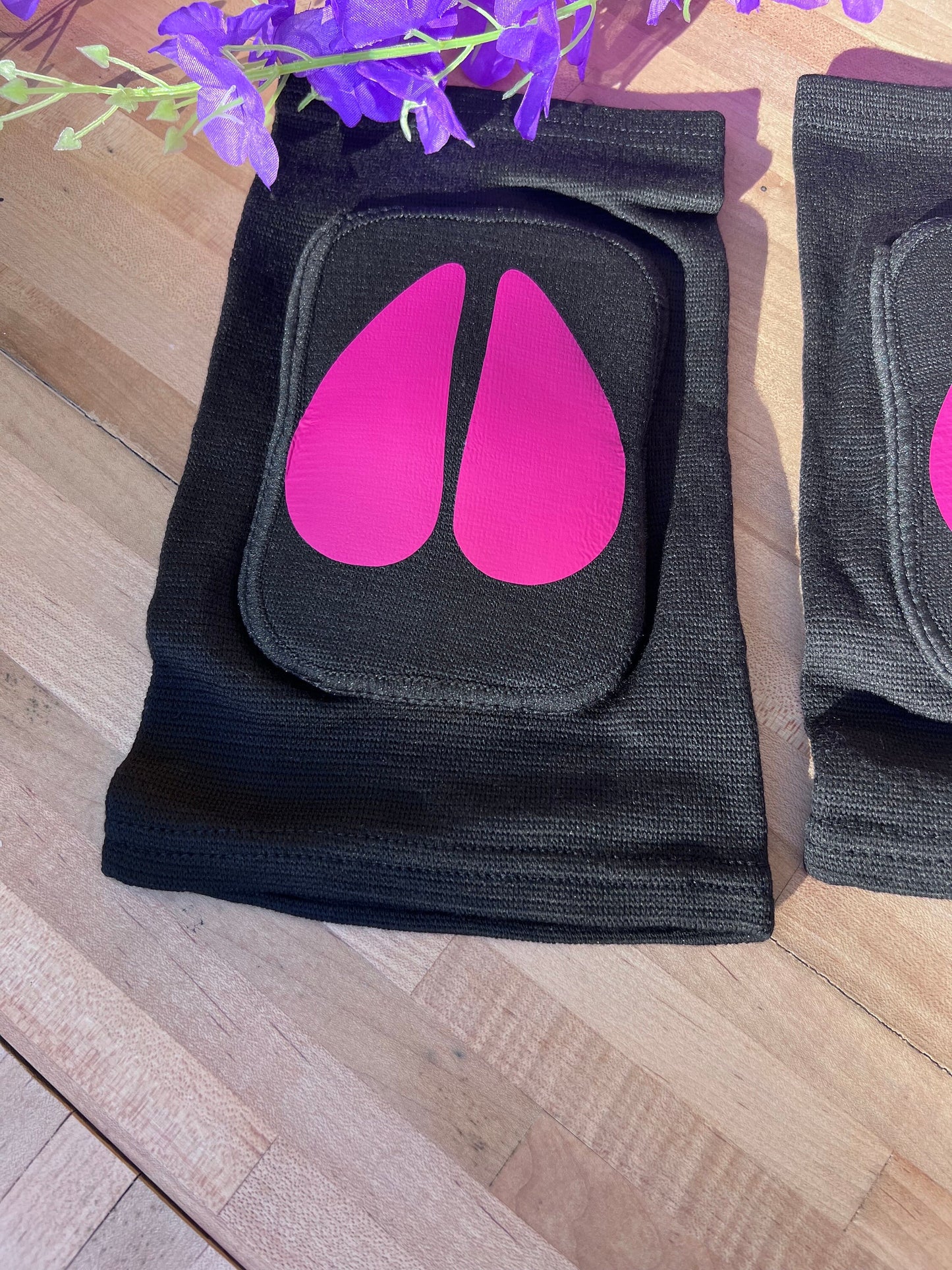 Another angle of the same black adult knee pads with hot pink cow hoof vinyl designs, shown on a wooden surface with a bunch of purple artificial flowers arranged above them.