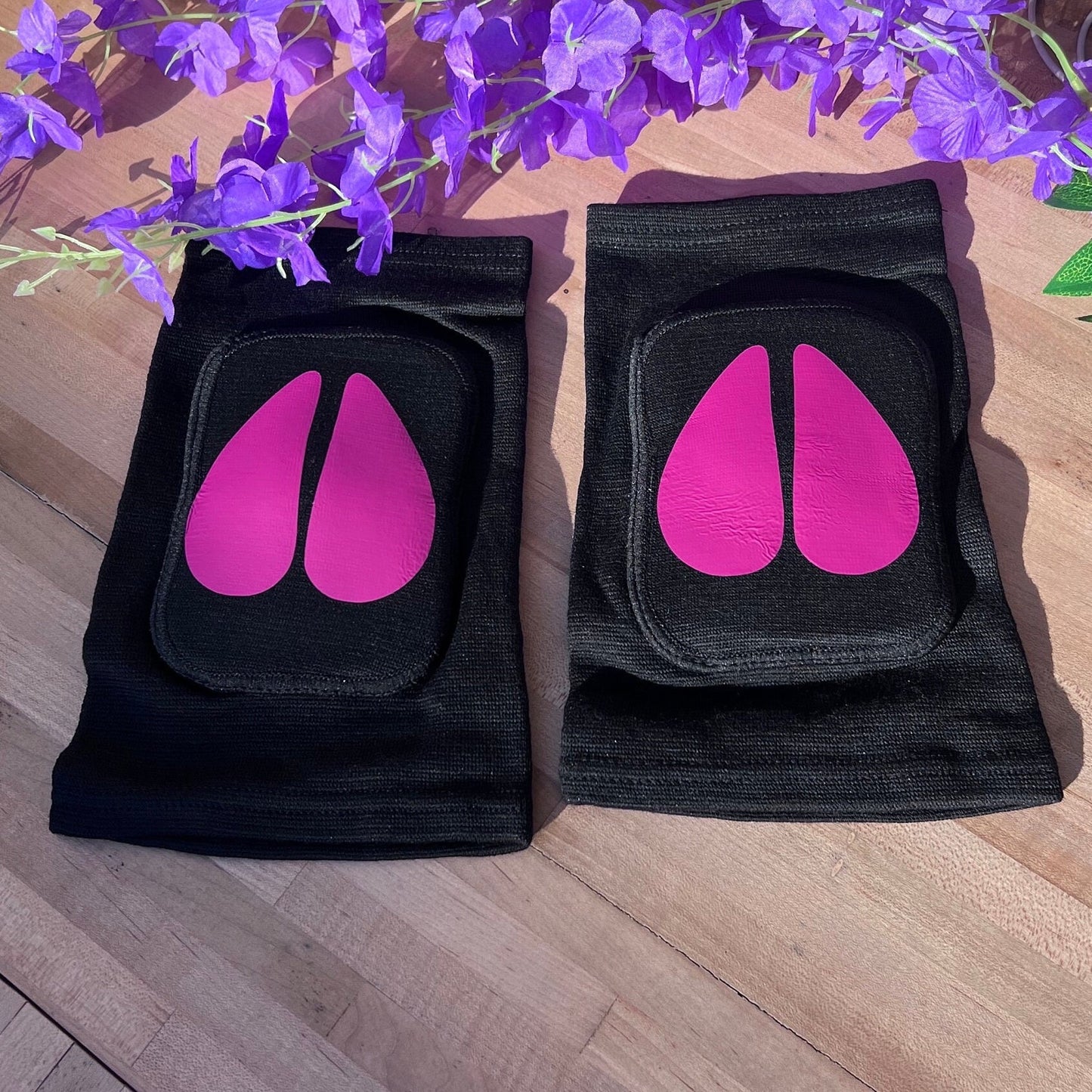 A different angle of the same black adult knee pads with bright pink cow hoof vinyl designs, shown on a wooden surface with a bunch of purple artificial flowers arranged above them.