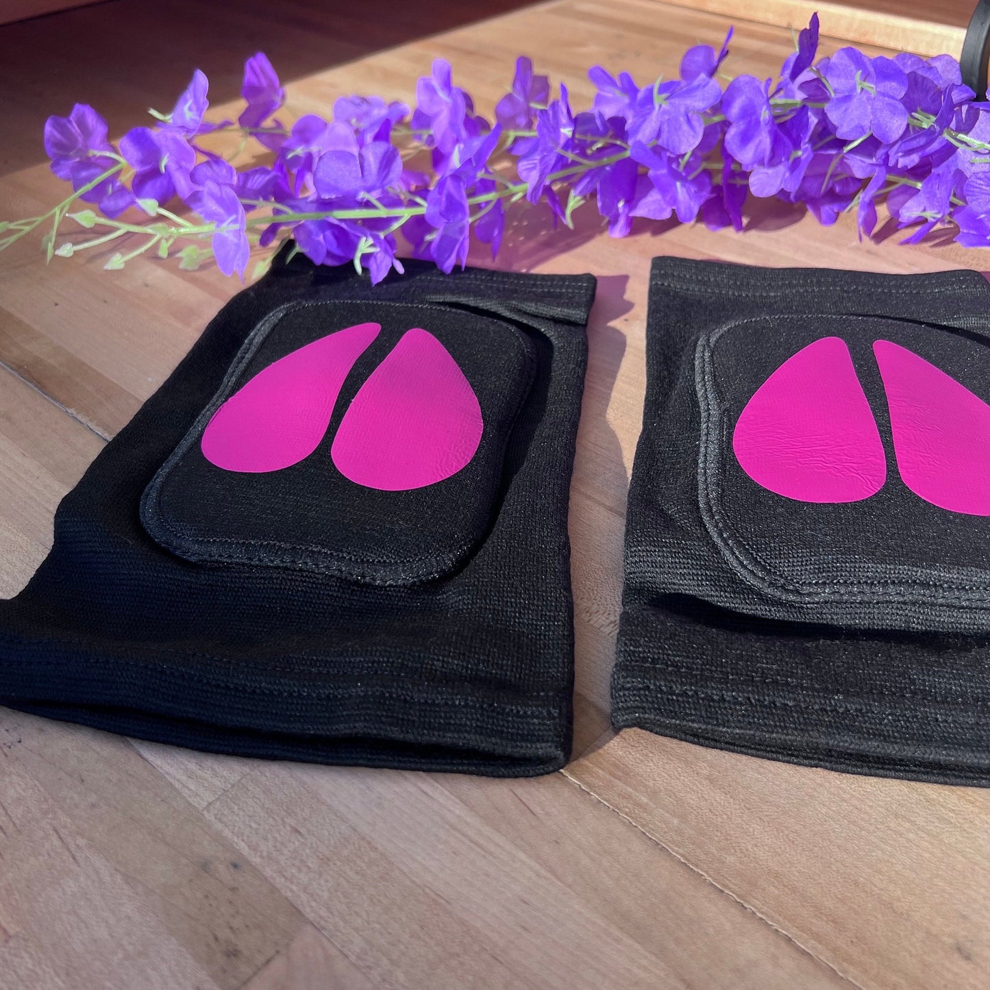 A pair of black adult knee pads with bright pink vinyl cow hoof designs in the center, placed on a wooden surface with purple artificial flowers in the background.