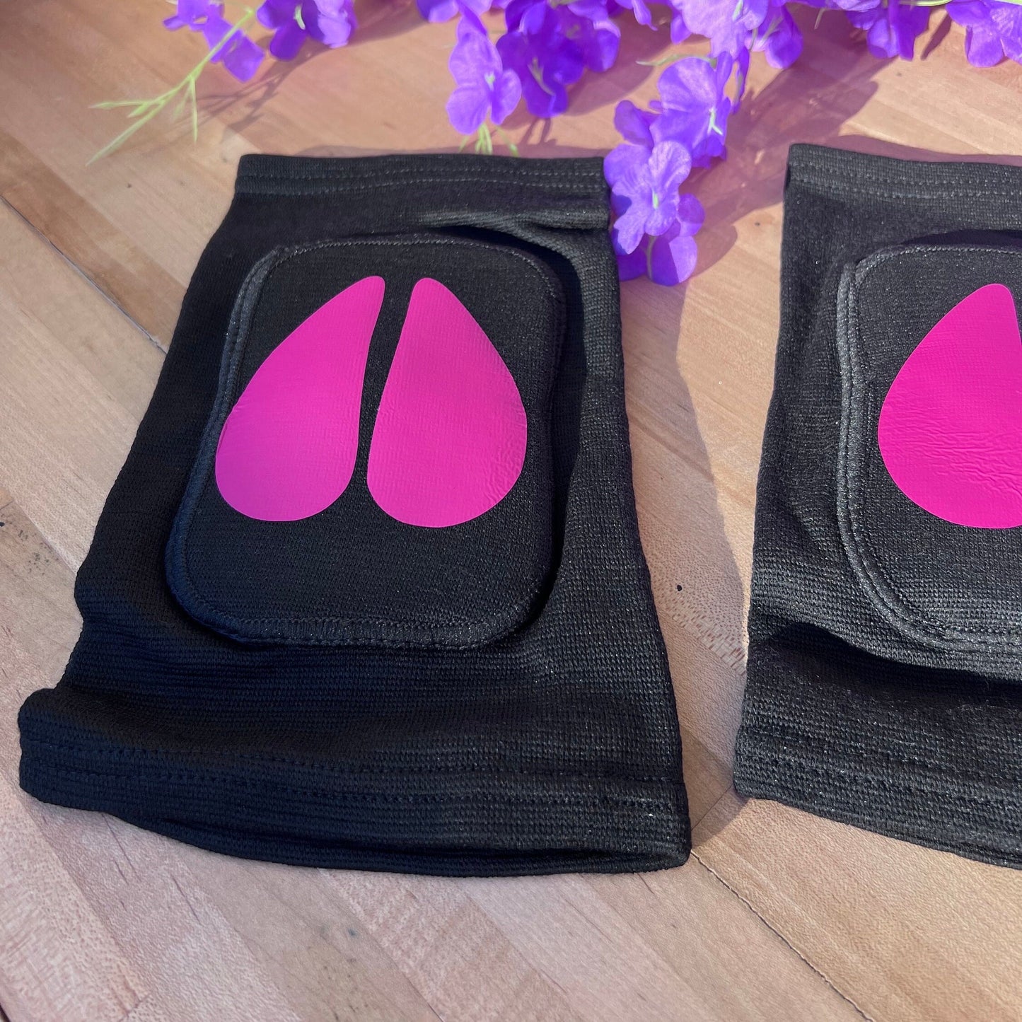 Close-up side view of black adult knee pads with hot pink cow hoof print, floral decor behind.