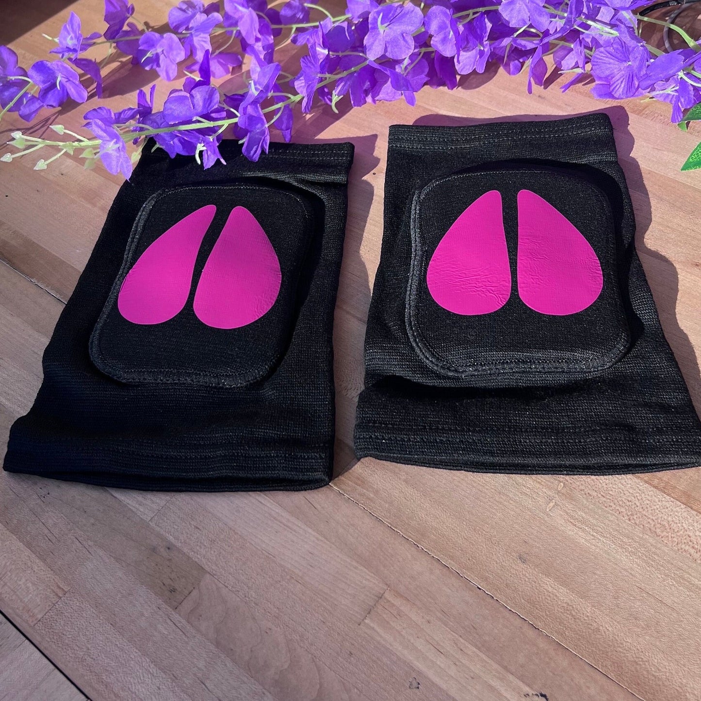 Another angle of the same black adult knee pads with bright pink cow hoof vinyl designs, shown on a wooden surface with a bunch of purple artificial flowers arranged above them.