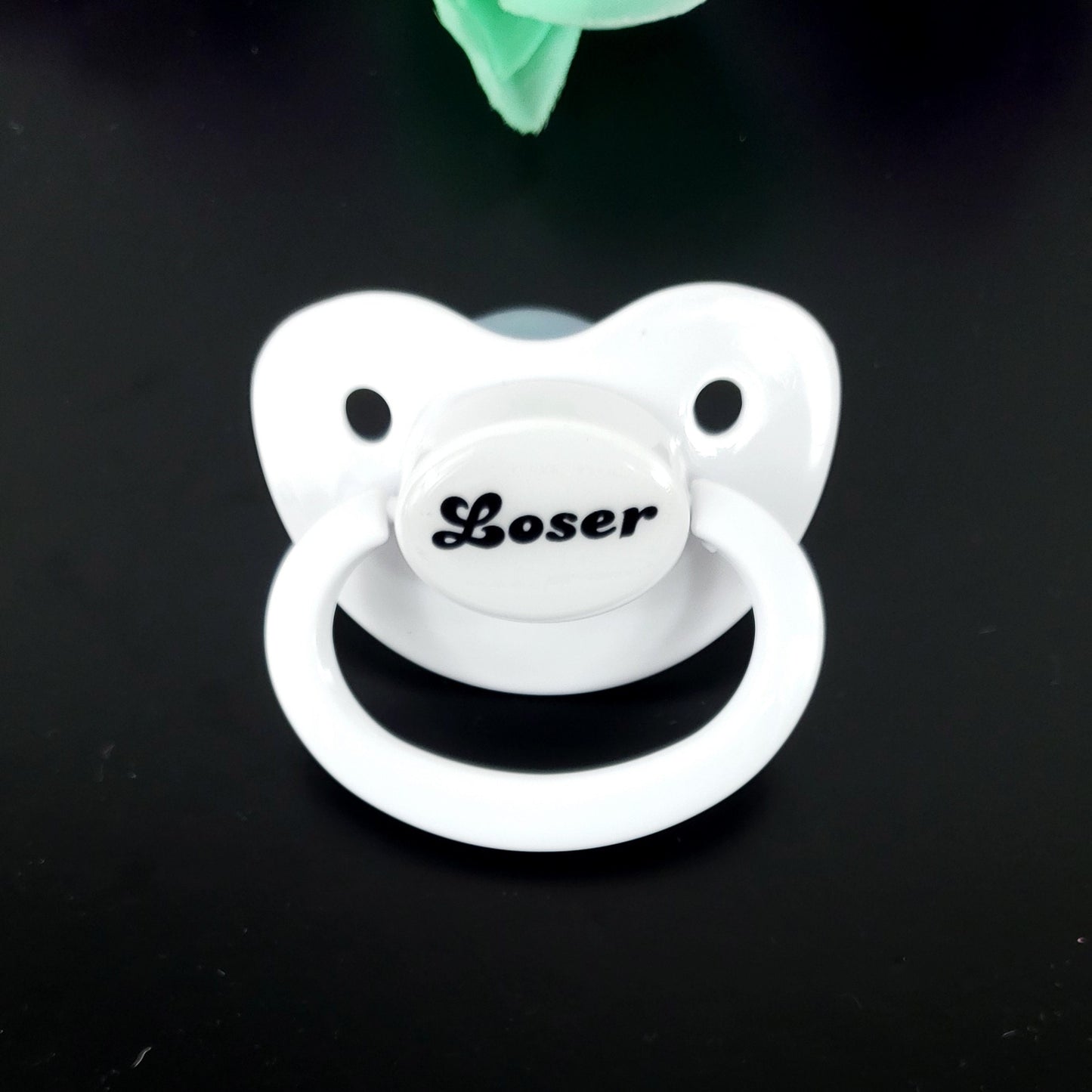 A white adult pacifier featuring bold black text in the center that reads "Loser," offering a simple and degrading design.