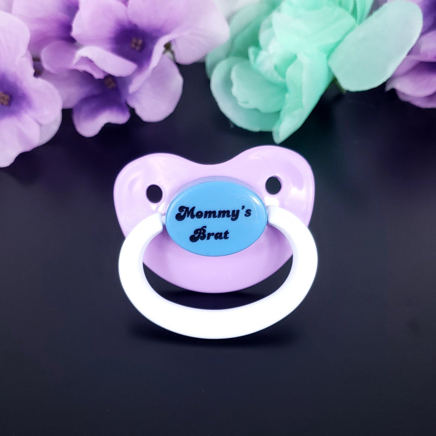 A pastel adult pacifier featuring bold white text that reads "Mommy's Brat" in the center, showcasing a playful and sassy design.