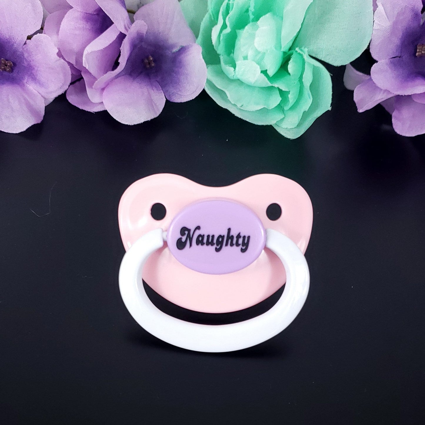 A pink and lavender adult pacifier with the text naughty on the center button, featuring a fun and playful design.