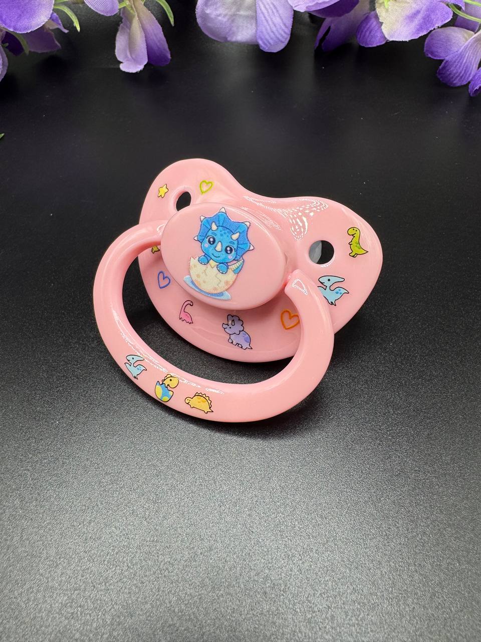 Baby pink adult pacifier with blue dinosaur coming out of it's shell on button with glow in the dark dinosaurs, hearts and stars