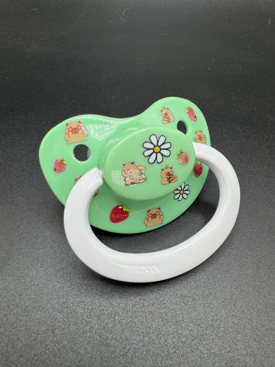 Pastel green adult pacifier with capybara, strawberry and flower designs on a dark gray background
