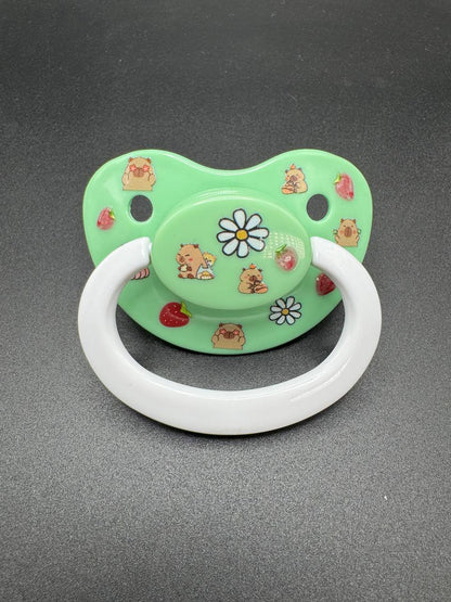 Green adult pacifier with capybara and flower designs on a gray background