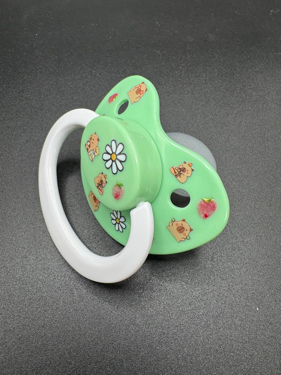 Green adult pacifier with white handle with strawberry capybara design on a dark gray background