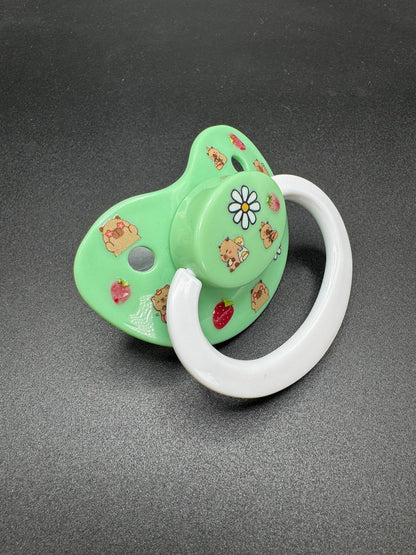 Green ABDL pacifier with cartoon capybara characters and flowers on it