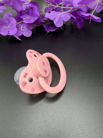 A pink pacifier decorated with small cherry illustrations, resting on a dark gray surface with purple flowers in the background.