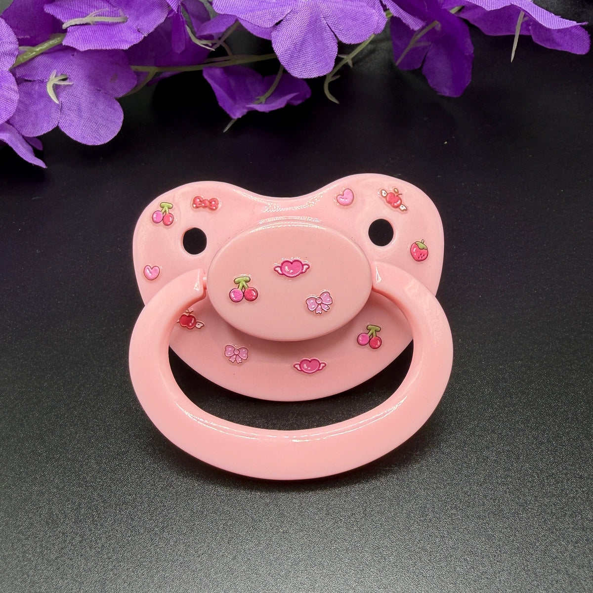An adult pink pacifier decorated with small cherry and flower motifs, displayed on a dark gray surface with purple flowers in the background.
