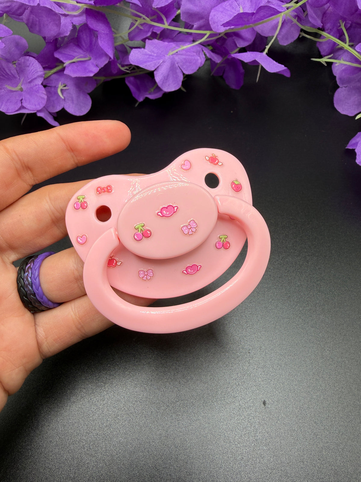 A pink adult pacifier decorated with small cherry and bow motifs, held in a hand against a dark background with purple flowers at the top.