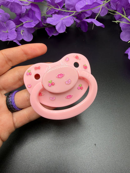 A pink adult pacifier decorated with small cherry and bow motifs, held in a hand against a dark background with purple flowers at the top.