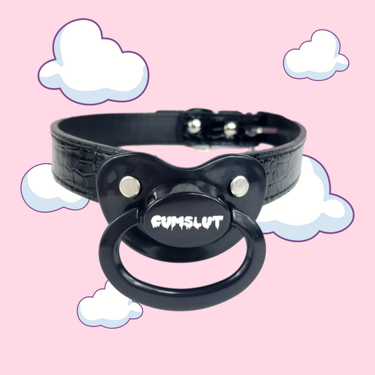 black adult pacifier with white text saying cumslut in the center. Attached to the pacifier is black adjustable straps.