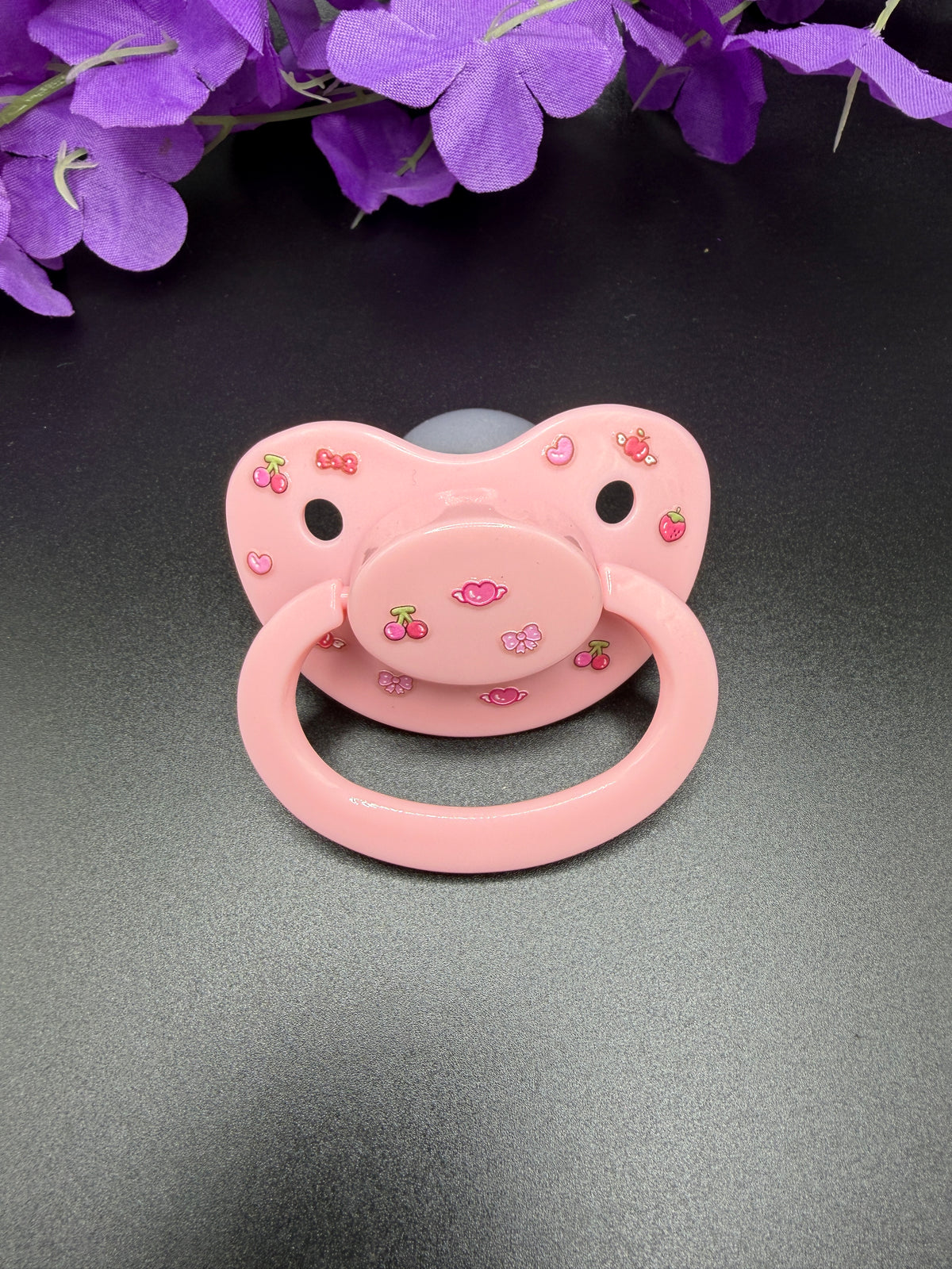 A pink silicone adult pacifier decorated with small cherry and strawberry motifs, displayed on a dark gray surface with purple flowers in the background.