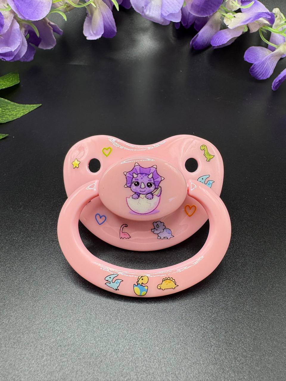 Baby pink adult pacifier with purple dinosaur coming out of egg on the button with glow in the dark dinosaurs and hearts on shield and handle