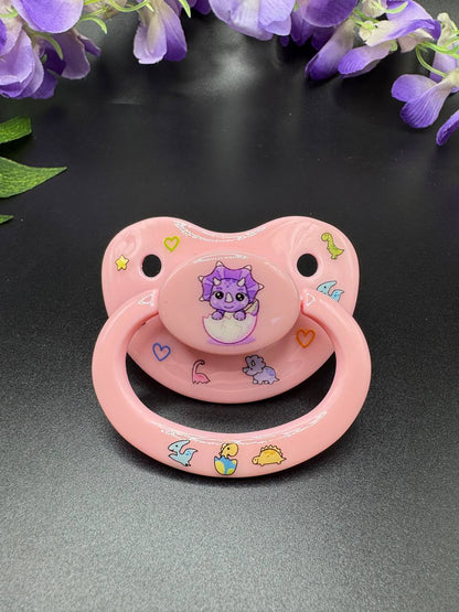 Baby pink adult pacifier with purple dinosaur coming out of egg on the button with glow in the dark dinosaurs and hearts on shield and handle