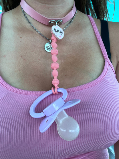 Person wearing a pink choker with a resin tag that reads "Kitty." A pink chewable pacifier clip is attached to the choker, ending in a lavender adult pacifier.