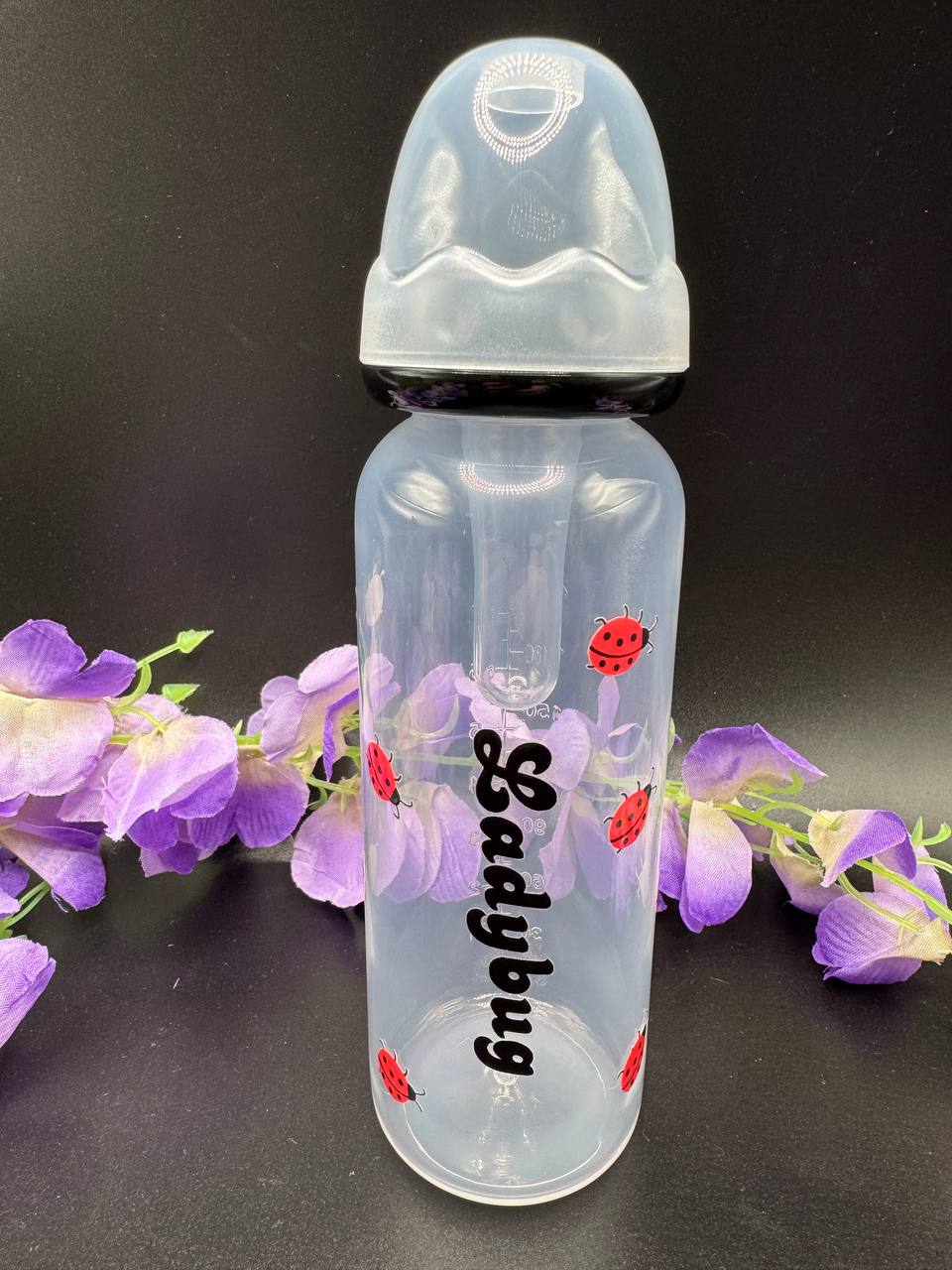 Transparent adult baby bottle with 'Ladybug' text and ladybug designs on a black background with purple flowers.