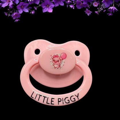 A pink adult pacifier with a cute pig graphic and the phrase 'LITTLE PIGGY' on it.